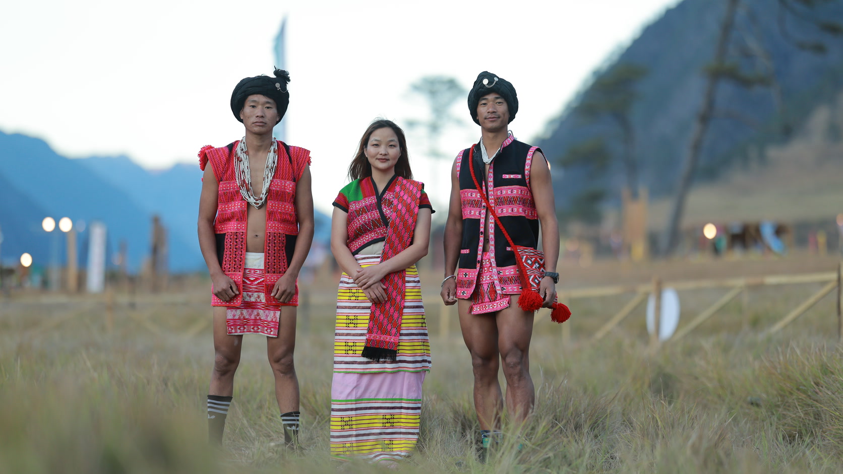 Three people in handwoven Arunachali dress standing in a meadow at dusk, a woman between two men, with mountain ridges and pine behind them